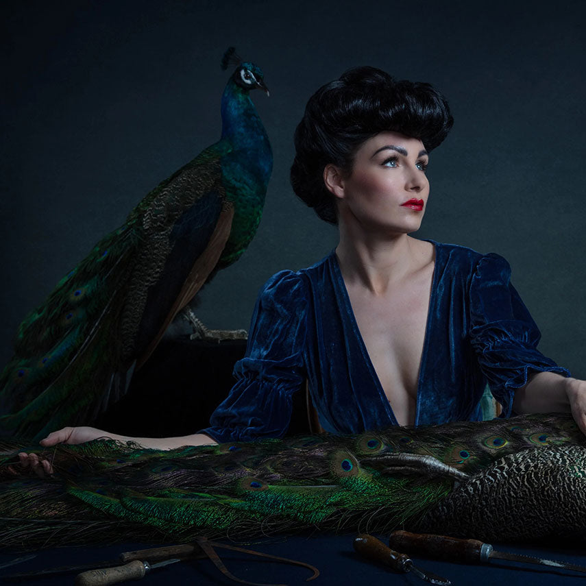 Allis Markham in a jewel toned blue dress, and a taxidermied peacock